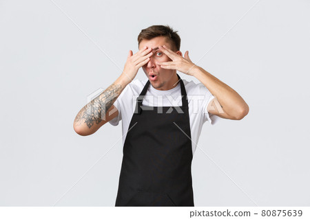 Employees, grocery stores and coffee shop concept. Intrigued funny barista, guy waiter in black apron, peeking through fingers as curious what happening, standing white background Employees, grocery stores and coffee shop concept. Intrigued funny barista, guy waiter in black apron, peeking through fingers as curious what happening, standing white background 80875639