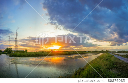 The pond reflects the light from the orange sun. The pool edge is green and yellow grass. The evening sky before the sunset is colorful. 80876085