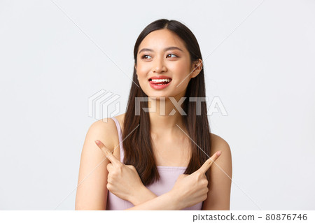 Beauty, fashion and people emotions concept. Dreamy happy beautiful asian woman in dress, smiling and lick lips from temptation, pointing fingers sideways two choices, white background 80876746