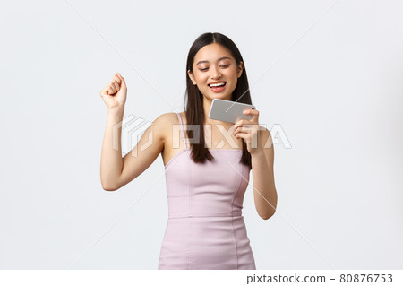 Luxury women, party and holidays concept. Happy satisfied pretty woman in evening dress, do champion dance as winning in mobile game, holding smartphone and fist pump excited, white background 80876753