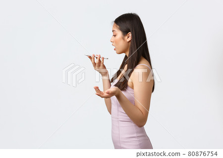 Luxury women, party and holidays concept. Profile of beautiful and stylish asian girl in evening dress record voice message, gesturing and holding mobile phone near mouth, white background Luxury women, party and holidays concept. Profile of beautiful and stylish asian girl in evening dress record voice message, gesturing and holding mobile phone near mouth, white background 80876754