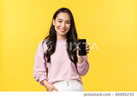 People emotions, lifestyle leisure and beauty concept. Smiling asian woman 20s, showing smartphone display, recommend application or mobile game, advertisement on screen, yellow background 80880230