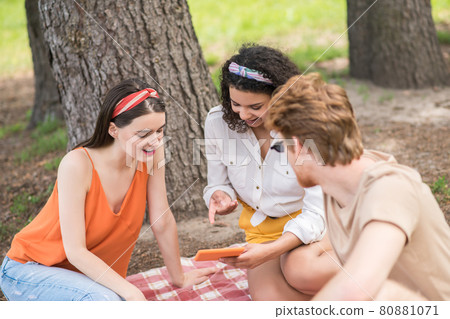 Two girls and guy with tablet at picnic Two girls and guy with tablet at picnic 80881071