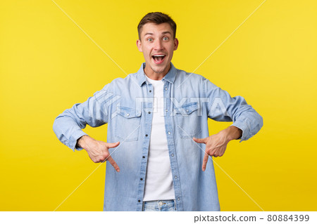 Enthusiastic handsome smiling man in casual outfit, pointing fingers down and looking amazed, telling big super good news, demonstrate advertisement, showing event promo, yellow background 80884399