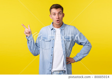 Curious and impressed blond handsome man, looking at camera excited while asking question about product, pointing upper left corner at interesting new special offer banner, yellow background 80885014