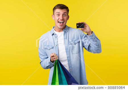 Shopping, leisure and advertisement concept. Enthusiastic handsome young man likes buying things, showing credit card and smiling, holding bags with purchased items, yellow background 80885064