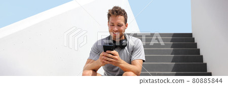 Young fit man using mobile phone app for online streaming of music listening with wireless headphones banner panoramic. City lifestyle. 80885844