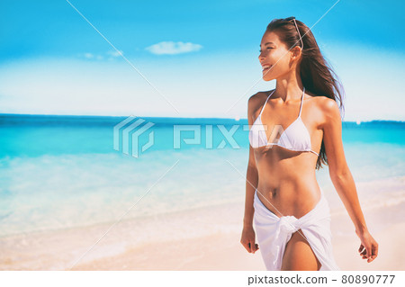 Bikini Asian woman in white swimsuit walking on ocean beach vacation for wellness spa luxury. Hair removal laser treatment summer ready body. Girl wearing pareo skirt beachwear. 80890777