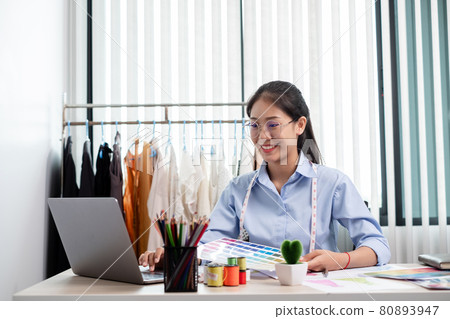 Beautiful young Asian fashion tailor with textile sewing accessories and entrepreneur designer sketches are full of bright colors on the desk with a laptop for creative ideas in the studio Beautiful young Asian fashion tailor with textile sewing accessories and entrepreneur designer sketches are full of bright colors on the desk with a laptop for creative ideas in the studio 80893947