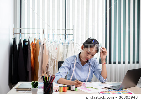 Beautiful young Asian fashion tailor with textile sewing accessories and entrepreneur designer sketches are full of bright colors on the desk with a laptop for creative ideas in the studio Beautiful young Asian fashion tailor with textile sewing accessories and entrepreneur designer sketches are full of bright colors on the desk with a laptop for creative ideas in the studio 80893948