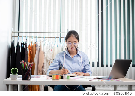 Beautiful young Asian fashion tailor with textile sewing accessories and entrepreneur designer sketches are full of bright colors on the desk with a laptop for creative ideas in the studio 80893949