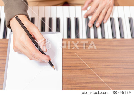 close up male composer hand writing a song on white blank music sheet while playing acoustic piano 80896139