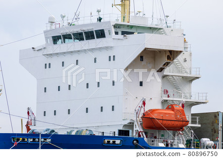 White bridge and orange enclosed lifeboat of a merchant ship under cargo handling 80896750