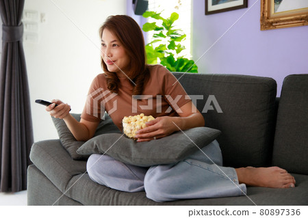 Mid adult Asian woman sitting on sofa in home living room holding bolw of popcorn and using remote control aiming to tv while watching in relax time Mid adult Asian woman sitting on sofa in home living room holding bolw of popcorn and using remote control aiming to tv while watching in relax time 80897336