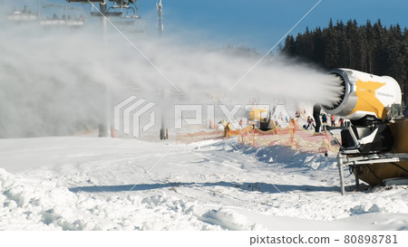Snow making system. Snow cannon makes artificial snow of water on sunny day in ski resort. On backdrop skiers slide down snow-covered white slope of mountain, ski lift working, winter forest, blue sky Snow making system. Snow cannon makes artificial snow of water on sunny day in ski resort. On backdrop skiers slide down snow-covered white slope of mountain, ski lift working, winter forest, blue sky 80898781