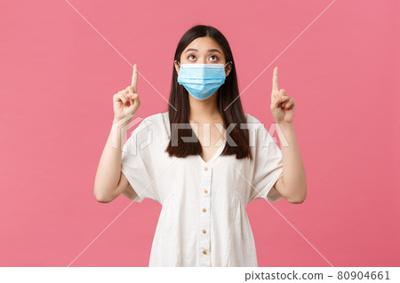 Covid-19, social distancing, virus and lifestyle concept. Curious pretty asian girl in medical mask and summer dress, looking and pointing fingers up at promo, advertisement, pink background 80904661