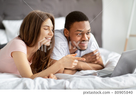 Cheerful millennial multinational couple lying on bed with laptop, watching fun video together Cheerful millennial multinational couple lying on bed with laptop, watching fun video together 80905840
