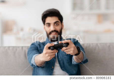 Weekend leisure. Joyful arab man using joystick and playing online video games, sitting on sofa in living room 80906181