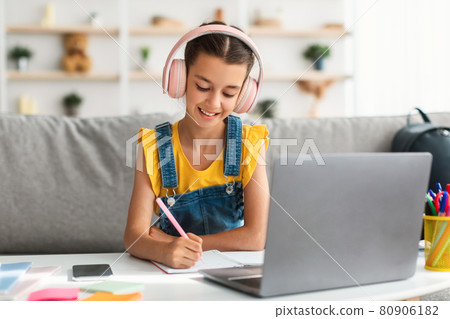 Smiling girl sitting at desk, using laptop, writing in copybook 80906182