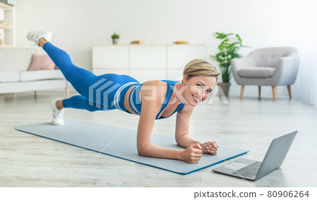 Happy woman doing plank on mat with laptop at home Happy woman doing plank on mat with laptop at home 80906264