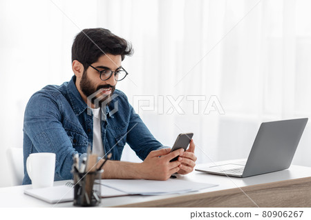 Arab freelancer guy using cellphone while working on laptop at desk at home office, browsing new app or messaging 80906267
