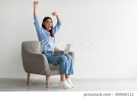 Full length of emotional woman with laptop raising hands, sitting in armchair, receiving great online news, white wall Full length of emotional woman with laptop raising hands, sitting in armchair, receiving great online news, white wall 80906312