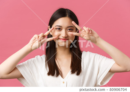 Beauty, people emotions and summer leisure concept. Cute and shy pretty japanese girl showing kawaii peace signs near eyes and smiling silly at camera, standing over pink background 80906704