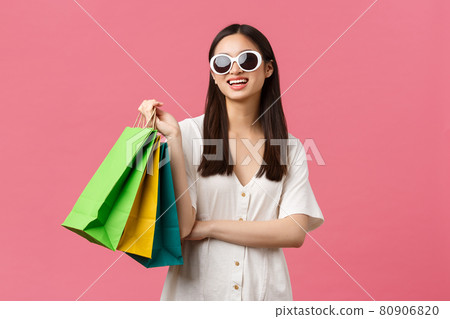 Beauty, people emotions and summer leisure and vacation concept. Carefree happy asian girl on vacation, tourist holding shopping bags and wearing sunglasses, smiling satisfied, pink background 80906820