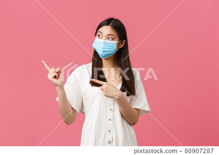 Covid-19, social distancing, virus and lifestyle concept. Curious cute asian girl, store visitor in medical mask asking question about product, pointing fingers left, standing pink background 80907287