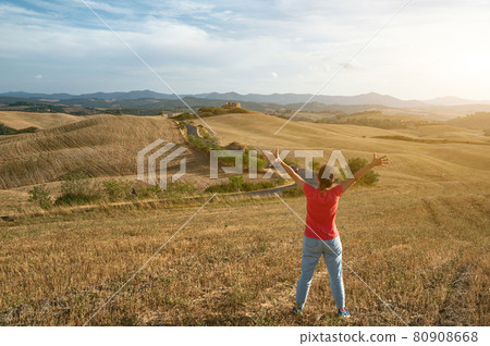 Volterra,Tuscany,Italy.August 2020.In the countryside of the city, a woman is inspired by nature. The landscape is crossed by the road. On her wrist the mask to protect against coronavirus,golden hour 80908668