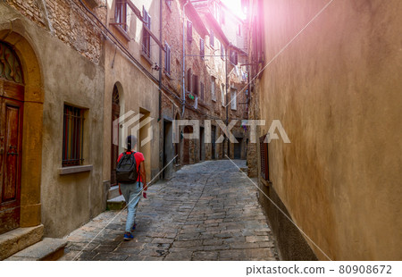 Volterra, Tuscany, Italy. Amazing shot of a charming alley in the historic center. A middle-aged tourist explores it. Sunbeams peek out at the end of the street. Volterra, Tuscany, Italy. Amazing shot of a charming alley in the historic center. A middle-aged tourist explores it. Sunbeams peek out at the end of the street. 80908672
