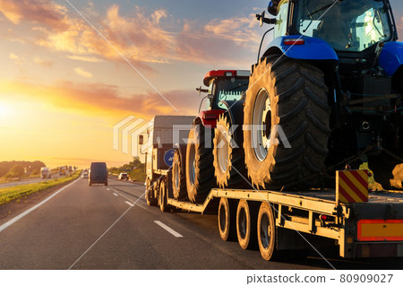 POV heavy industrial truck semi trailer flatbed platform transport two big modern farming tractor machine on common highway road at sunset sunrise sky. Agricultural equipment transportation service 80909027