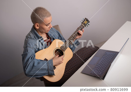 Young caucasian woman with short blonde hair playing guitar and watching training video on laptop. 80911699