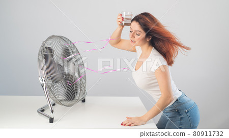 A red-haired Caucasian woman chills by the electric fan and drinks a cold drink. Climate control in the apartment 80911732