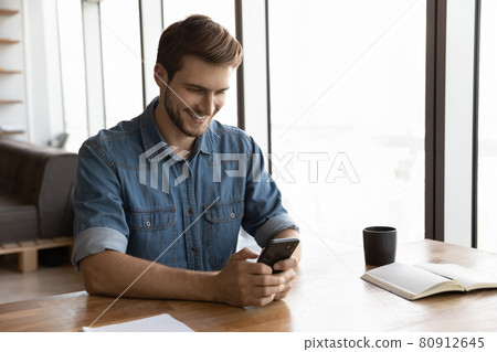 Happy young millennial man with smartphone sitting at table Happy young millennial man with smartphone sitting at table 80912645