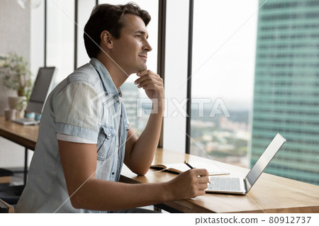 Thoughtful happy young millennial man working at laptop in apartment Thoughtful happy young millennial man working at laptop in apartment 80912737