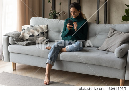 Full length smiling young woman using phone, relaxing on couch Full length smiling young woman using phone, relaxing on couch 80913015