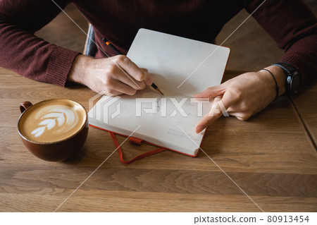 A young man sits in cafe, drinks coffee and makes notes and sketches in notebook. A young man sits in cafe, drinks coffee and makes notes and sketches in notebook. 80913454