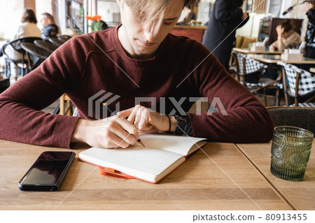 A young man sits in cafe, drinks coffee and makes notes and sketches in notebook. A young man sits in cafe, drinks coffee and makes notes and sketches in notebook. 80913455