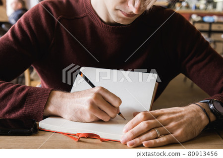 A young man sits in cafe, drinks coffee and makes notes and sketches in notebook. A young man sits in cafe, drinks coffee and makes notes and sketches in notebook. 80913456