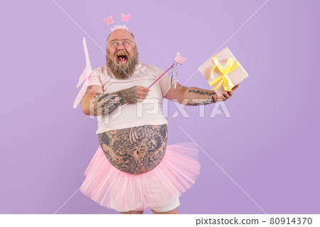 Excited obese male person in fairy costume holds gift box on purple background Excited obese male person in fairy costume holds gift box on purple background 80914370