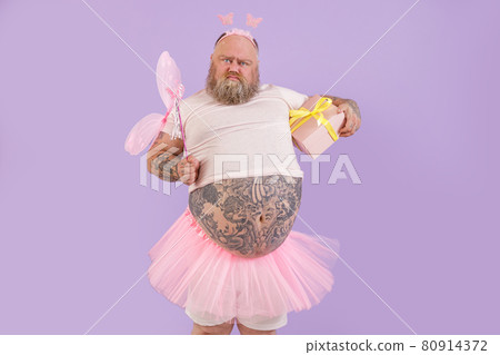Funny bearded plump man in fairy costume with gift box grimaces on purple background Funny bearded plump man in fairy costume with gift box grimaces on purple background 80914372