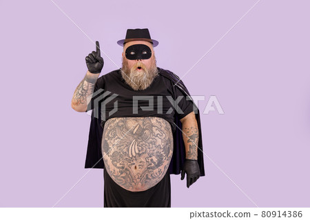 Surprised man with overweight in hero suit got idea and points up on purple background Surprised man with overweight in hero suit got idea and points up on purple background 80914386