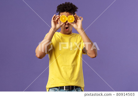 Holidays, vitamins and vacation concept. Portrait of funny, playful young guy fool around, playing with fruits, making eyes from pieces of orange, show wondered face expression, purple background 80914503