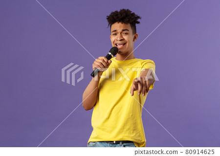 This song is for you. Portrait of romantic carefree hispanic man singing karaoke, pointing at camera as dedicate his performance, holding microphone, standing purple background 80914625