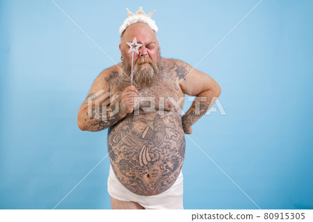 Tattooed obese man with crown looks through star on magic stick on blue background 80915305