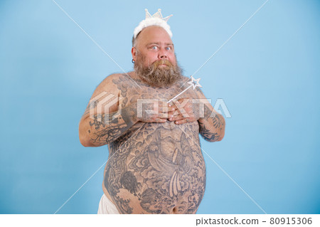 Funny bearded man with overweight wearing toy crown covers breasts on light blue background Funny bearded man with overweight wearing toy crown covers breasts on light blue background 80915306