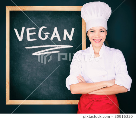 Vegan menu woman chef wearing uniform showing blackboard sign at restaurant for today's special. Vegeterian food. Word VEGAN handwritten on black chalkboard advert concept Vegan menu woman chef wearing uniform showing blackboard sign at restaurant for today's special. Vegeterian food. Word VEGAN handwritten on black chalkboard advert concept 80916330