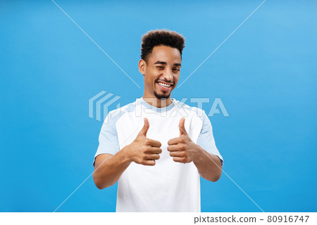 You doing great, support you. Satisfied creative and stylish young african american man with beard winking joyfully smiling broadly showing thumbs up in approval or like gesture over blue wall 80916747