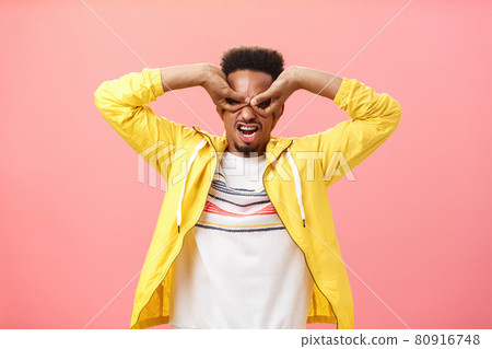 Man from childhood dreamt become superhero. Childish playful african american young guy acting immature making mask from hands over eyes aping standing in yellow jacket over pink wall 80916748